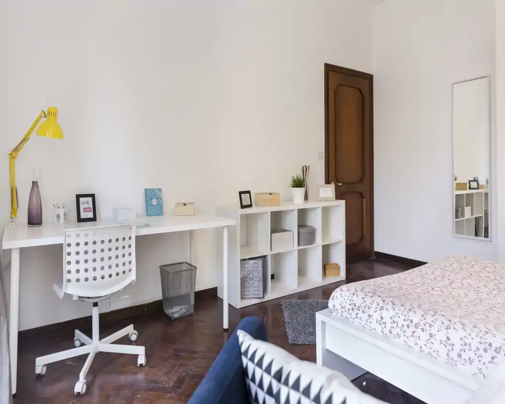 Camera da letto luminosa e moderna con struttura del letto bianca e copriletto a fiori, una postazione di lavoro con scrivania lunga e scaffalatura e pavimenti in legno caldi. Ambiente pulito, minimal e dal tocco nordico con pratici spazi contenitivi.