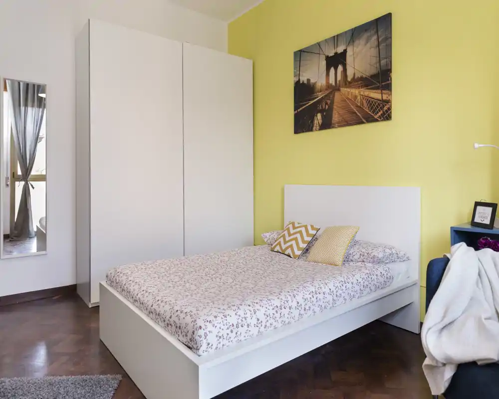 Camera da letto moderna e luminosa con letto matrimoniale bianco, biancheria a fantasia e parete d'accento gialla; armadio e specchio offrono spazio e riflesso di luce naturale.