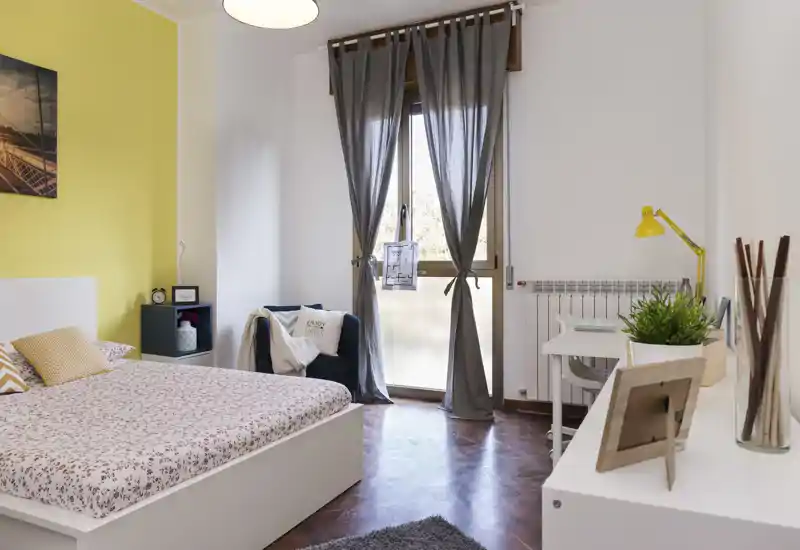 Camera da letto luminosa e curata con struttura del letto bianca, biancheria a fiori e tessili morbidi; la luce naturale dalla finestra alta e gli arredi armoniosi creano un ambiente accogliente.