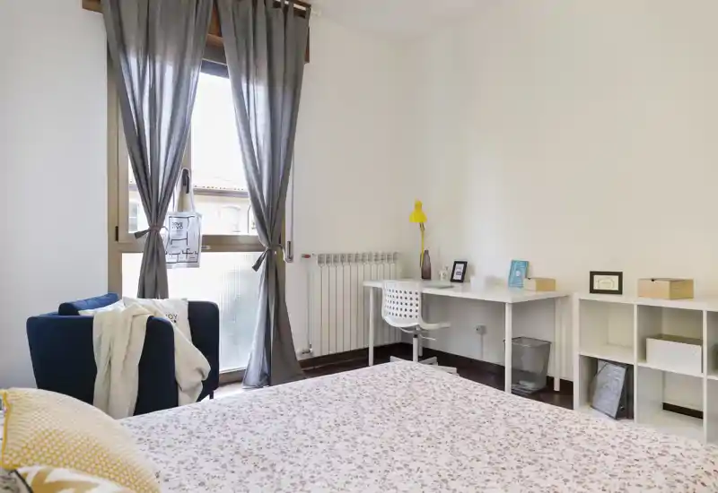 Camera da letto luminosa e ben arredata con copriletto floreale, una comoda poltrona vicino alla finestra e una scrivania minimal: ideale per riposo e lavoro.