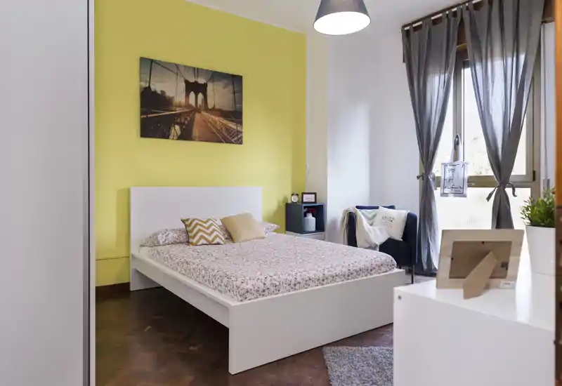 Camera da letto luminosa e di stile con letto a piattaforma bianco su una parete gialla, cuscini decorativi e quadro appeso. La luce naturale dalla finestra con tenda e i tessuti creano un'atmosfera accogliente.