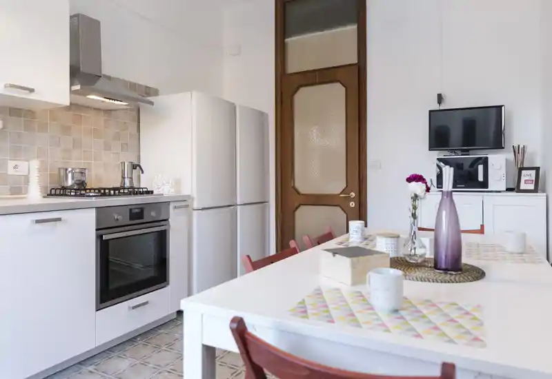 Bright modern kitchen with white cabinets, gas stove and fridge, plus a dining table set with colorful placemats and a decorative vase — ideal for apartment listings.