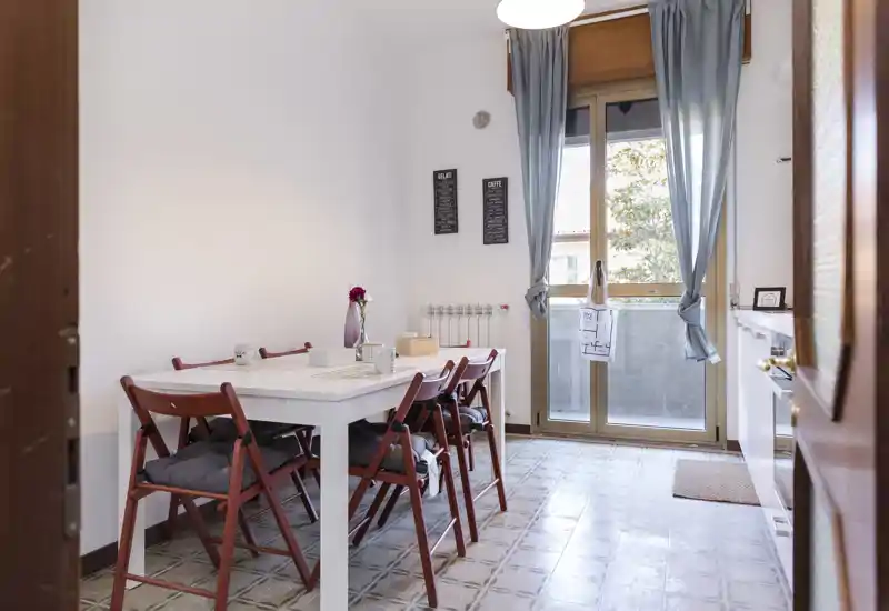 Bright, furnished dining area with a white table, four wooden folding chairs with cushions and a large window/door letting in natural light; simple, homely styling ideal for meals.