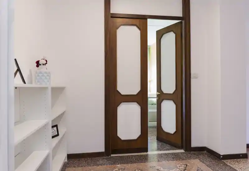 Bright entrance/hallway with decorative terrazzo floor, a wooden doorway leading to an adjoining room and a white shelving unit for storage.