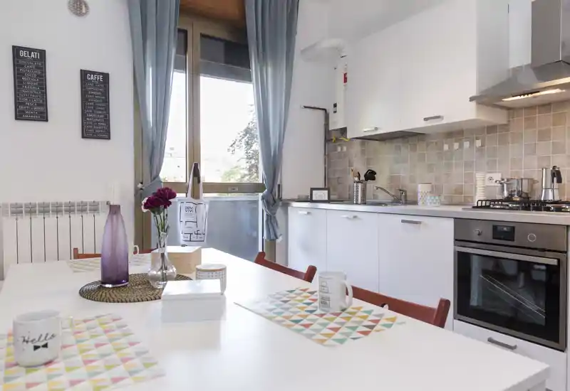 Bright, modern kitchen with white cabinets, gas stove and oven, and a dining table set with colorful placemats and mugs. Large window provides natural light and the styled details (vase, textiles) create a welcoming atmosphere.