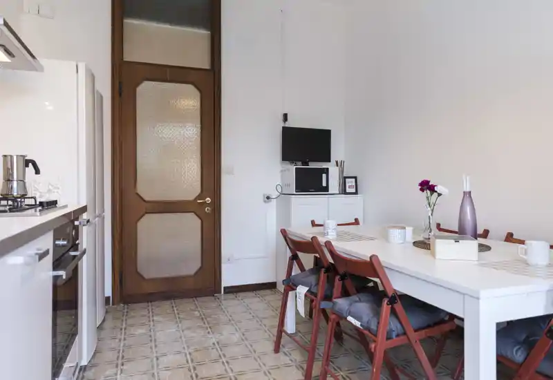Bright, tidy kitchen with an integrated dining area featuring a white table, wooden folding chairs with cushions, and modern appliances along the left wall. Decorative touches like a vase with flowers and placemats make the space welcoming for meal occasions.