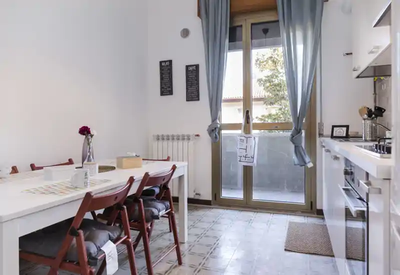 Bright, modern kitchen with a dining table for four next to a large window/door that opens to a small balcony. Clean white cabinetry, patterned floor tiles and cozy chair cushions create a welcoming cooking and dining area.