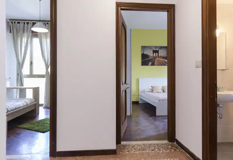 Bright, simply styled bedroom visible through a doorway, featuring a white bedframe, patterned pillows and a framed wall print; soft natural light and warm wooden floors create a welcoming feel.