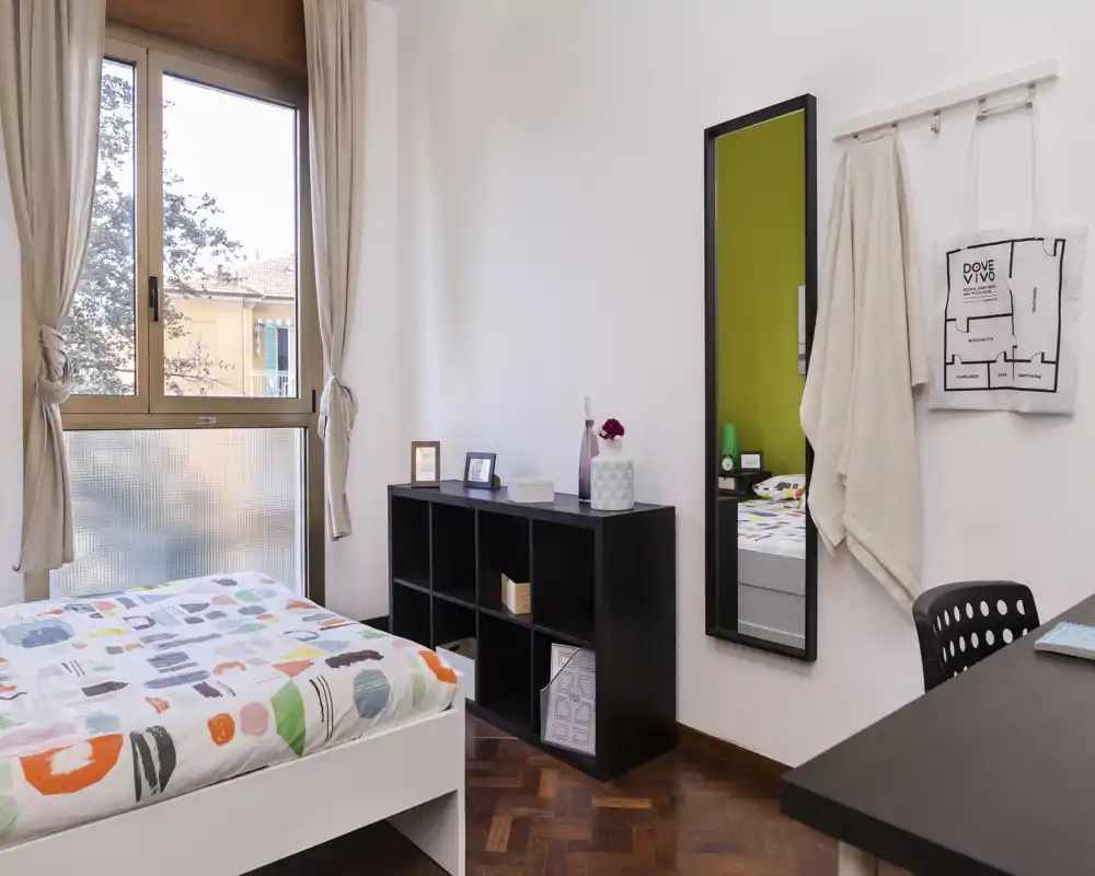Bright, neatly staged single bedroom with a patterned duvet, large window providing natural light, a shelving unit and a desk — ideal for a comfortable rental.