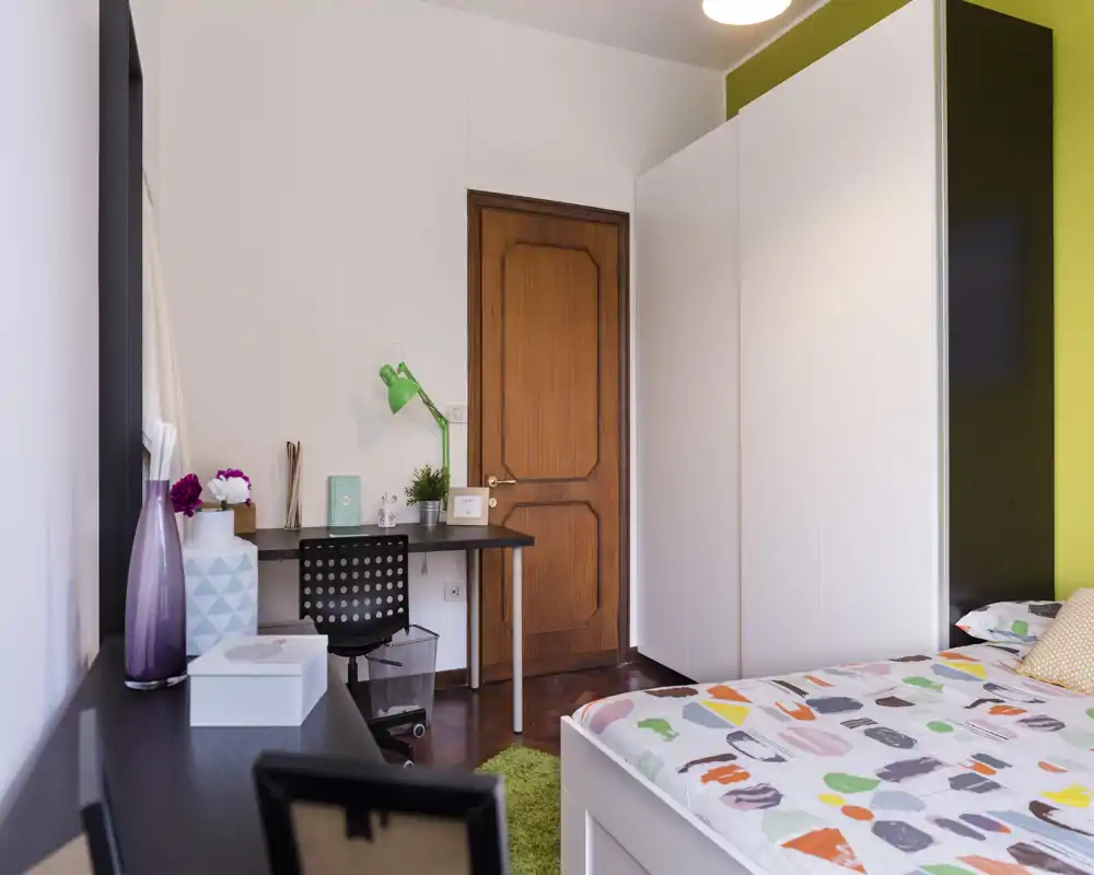 Bright, modern bedroom with patterned bed linens, a large white wardrobe and a compact workspace with green desk lamp — ideal for students or young professionals.
