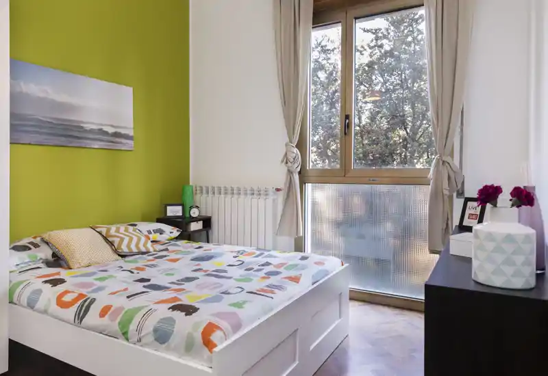 Dormitorio moderno y luminoso con cama blanca, edredón colorido y cojines, y una gran ventana que aporta luz natural. Cortinas neutras y decoración sencilla crean un ambiente acogedor.