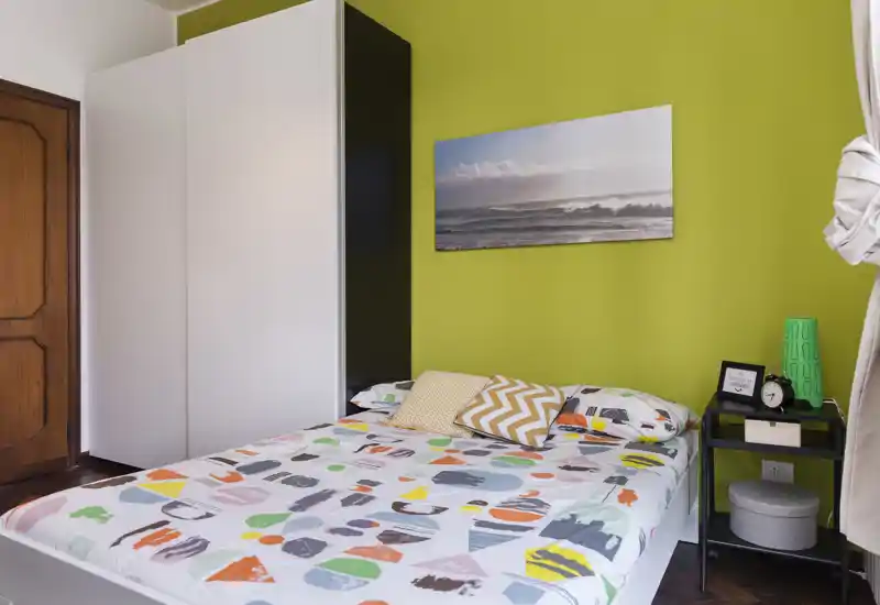 Dormitorio luminoso y colorido con cama doble, cojines decorativos, mesita y armario frente a una pared verde; estilo ordenado y luz natural ideal para anuncios de alquiler.