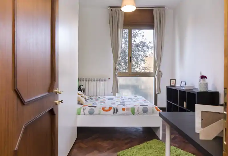 Dormitorio luminoso y bien presentado con cama doble frente a una gran ventana. Paredes neutras y suelos de madera crean una sensación de calma; estantería y detalles decorativos hacen el espacio acogedor.