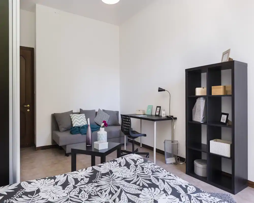 Dormitorio luminoso y bien preparado con cama estampada en primer plano, sofá compacto, escritorio y estantería — ideal para una persona o pareja que busque un espacio funcional y acogedor.