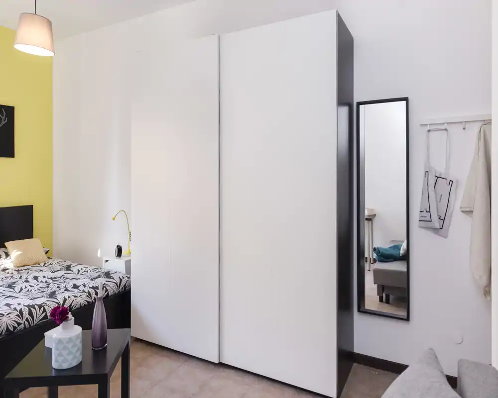 Dormitorio moderno y luminoso con cama estampada, mesita y gran armario blanco. Espacio limpio y bien iluminado con detalles decorativos sencillos y espejo de cuerpo entero.