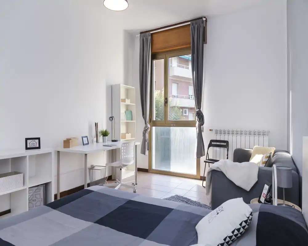 Chambre moderne et lumineuse avec grande fenêtre, lit double au linge à motifs, bureau et étagères pour travailler, et un petit coin salon près de la fenêtre.