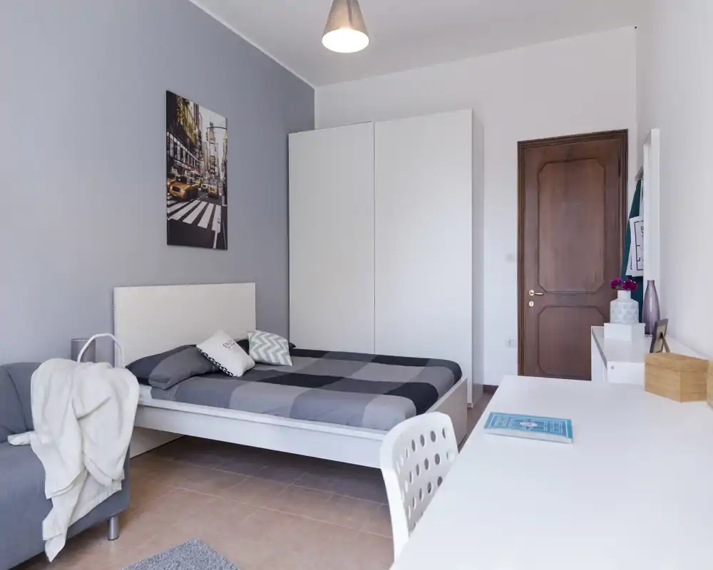 Chambre moderne et lumineuse avec lit double, armoire blanche et espace de travail. Palette grise et blanche, décoration soignée et lumière naturelle pour une ambiance calme et accueillante.