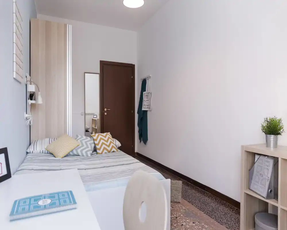 Chambre simple claire et soignée avec lit fait, coussins à motifs et un petit bureau au premier plan; étagère minimaliste et miroir près de la porte donnent une impression d'espace.