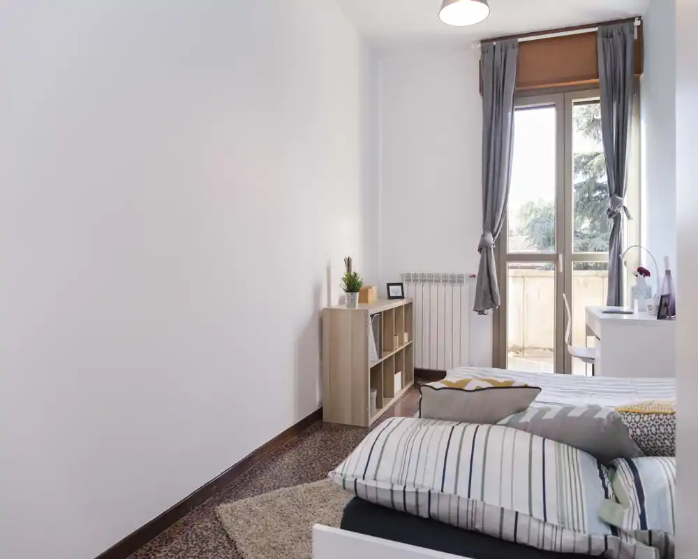 Chambre lumineuse et sobre avec lit double, coussins et petite étagère près d'une fenêtre donnant sur un balcon apportant de la lumière naturelle.