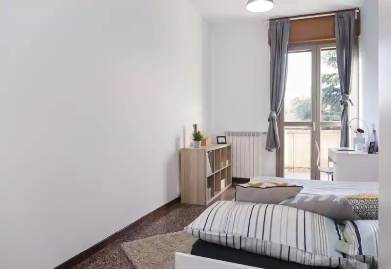 Bright, simply styled bedroom with a double bed, cushions and a small shelving unit next to a balcony window letting in natural light.
