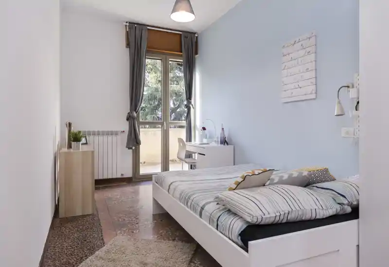 Chambre lumineuse et soignée avec lit double, linge de lit rayé, lampe de chevet et petit bureau près de la porte du balcon. Mur bleu clair et lumière naturelle offrent une ambiance apaisante.