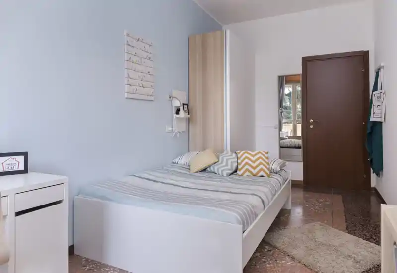 Chambre lumineuse et bien mise en scène avec un lit blanc complet, coussins tendance et un tapis doux ; mur pastel et lumière naturelle créent une ambiance chaleureuse.