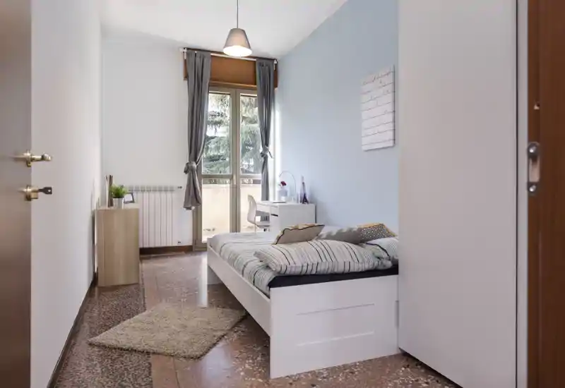 Chambre simple lumineuse et soignée avec un lit blanc, une literie rayée, un petit bureau près du balcon et une belle lumière naturelle.