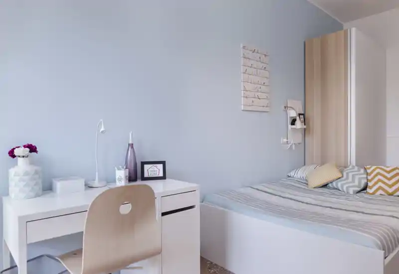 Chambre moderne et lumineuse avec un lit simple bien fait, linge de lit rayé et coussins décoratifs; un petit bureau blanc et une chaise offrent un coin travail soigné sous une lumière douce.