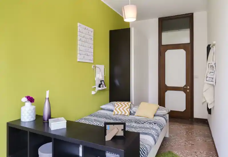 Chambre lumineuse et moderne avec lit double à motifs, coussins décoratifs et mur d'accent vert. La pièce dispose d'une porte en bois, d'étagères près du lit et d'un meuble bas au pied du lit.