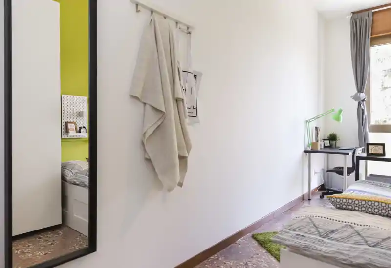 Coin chambre lumineux et moderne avec un lit simple, une literie à motifs, un petit bureau avec une lampe verte près de la fenêtre et une grande glace murale.