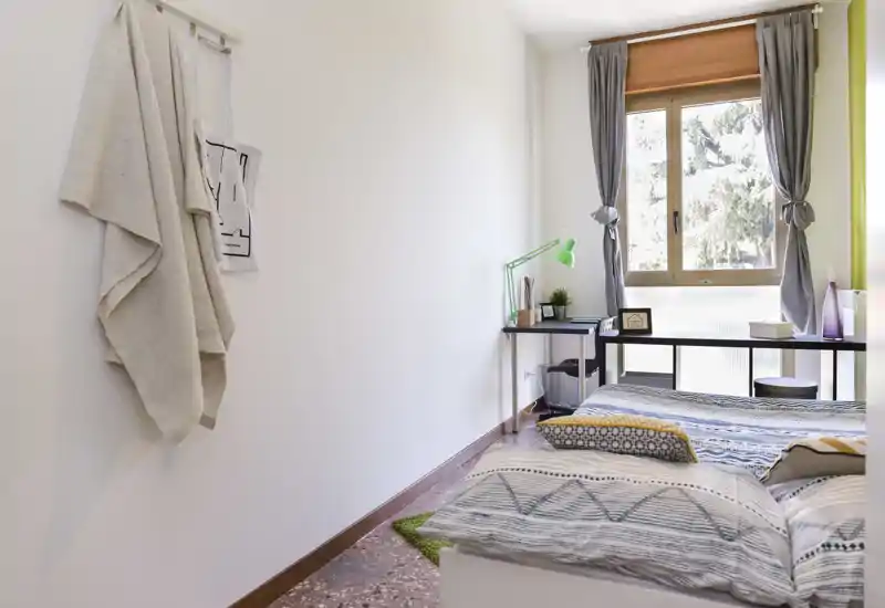 Chambre lumineuse et soignée avec literie à motifs, lit bas près d’une grande fenêtre et un bureau étroit avec lampe verte — parfaite pour une chambre simple ou d’appoint.