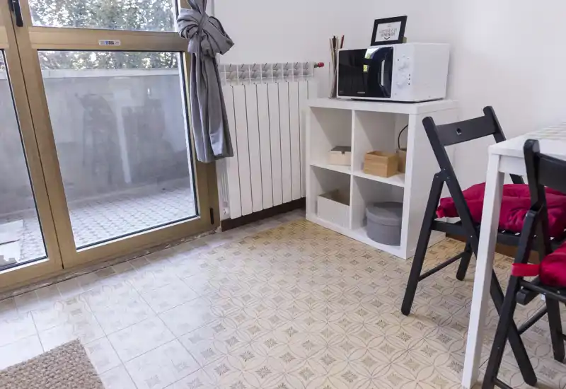 Small, bright kitchen corner with a large patio door, white storage cube and microwave; simple dining area with folding chairs and colorful cushions.