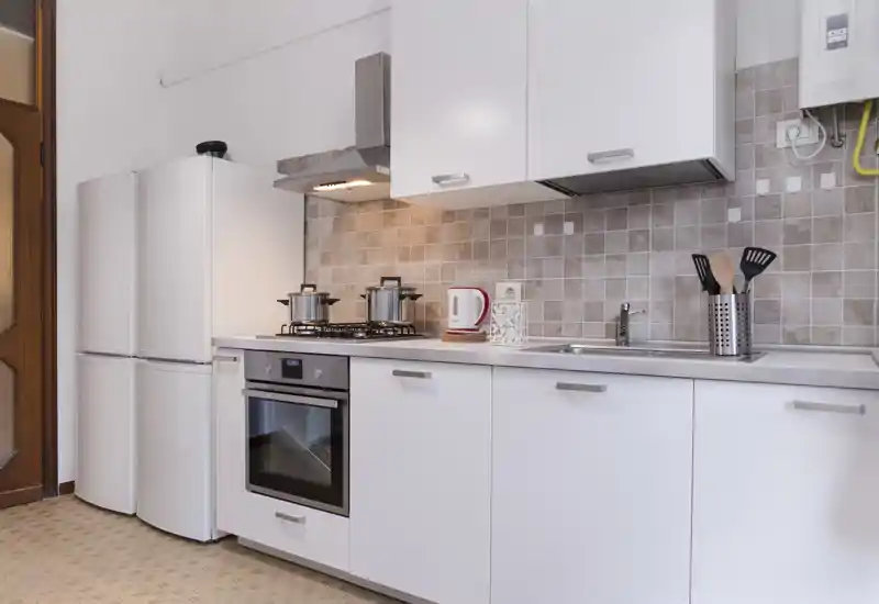 Modern, compact kitchen with white cabinets, built-in oven, gas hob and fridge. Neutral tiled backsplash and neat countertop with kettle and utensils; well lit and ready for cooking.