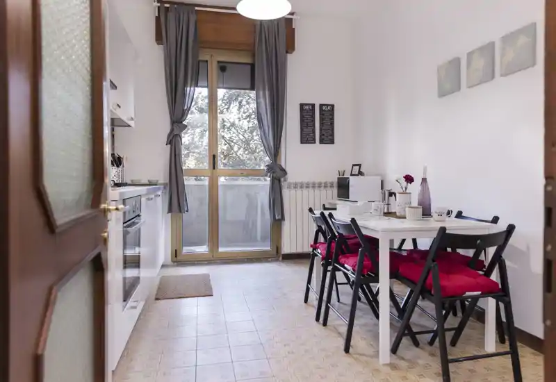 Bright, neatly staged kitchen with a dining table for four. Natural light from the balcony door, modern appliances along the left wall, and red seat cushions add a warm, inviting touch.