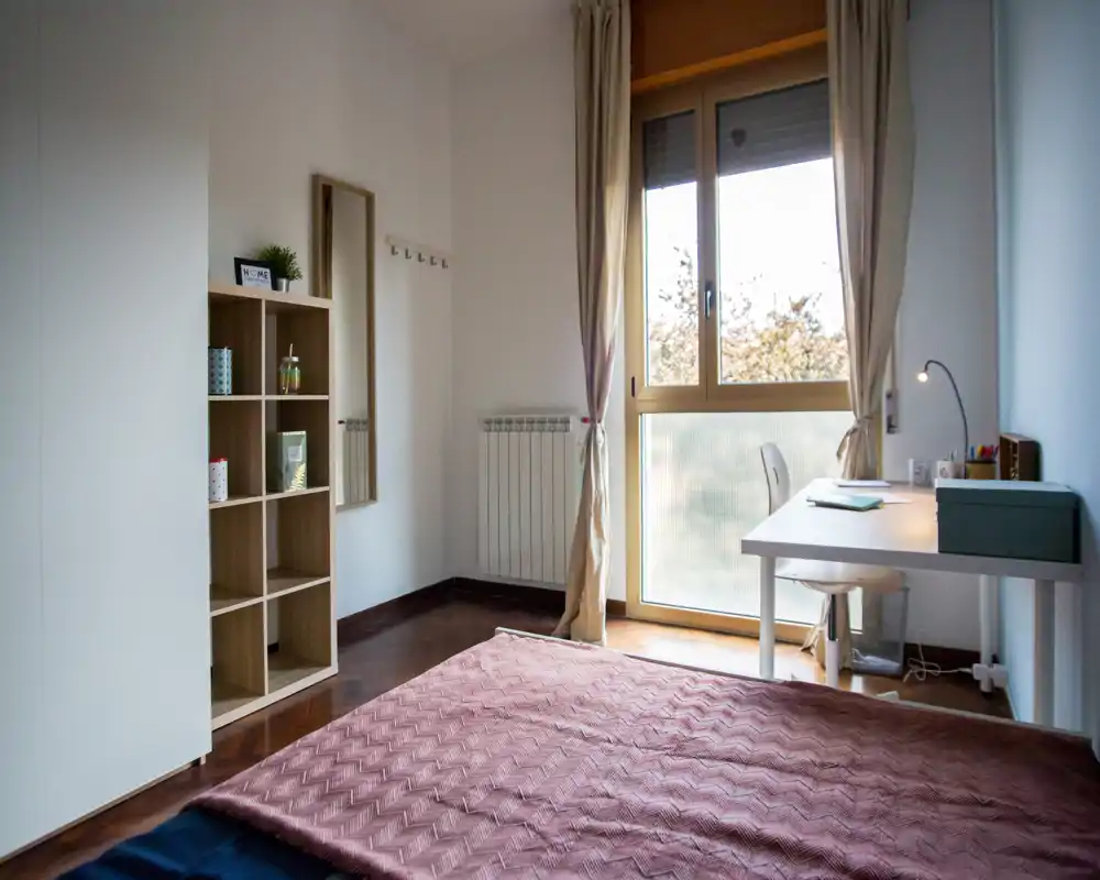 Camera da letto luminosa e arredata in modo semplice con letto matrimoniale in primo piano, scrivania vicino alla finestra e scaffalatura per riporre oggetti; ampia finestra con luce naturale gradevole.