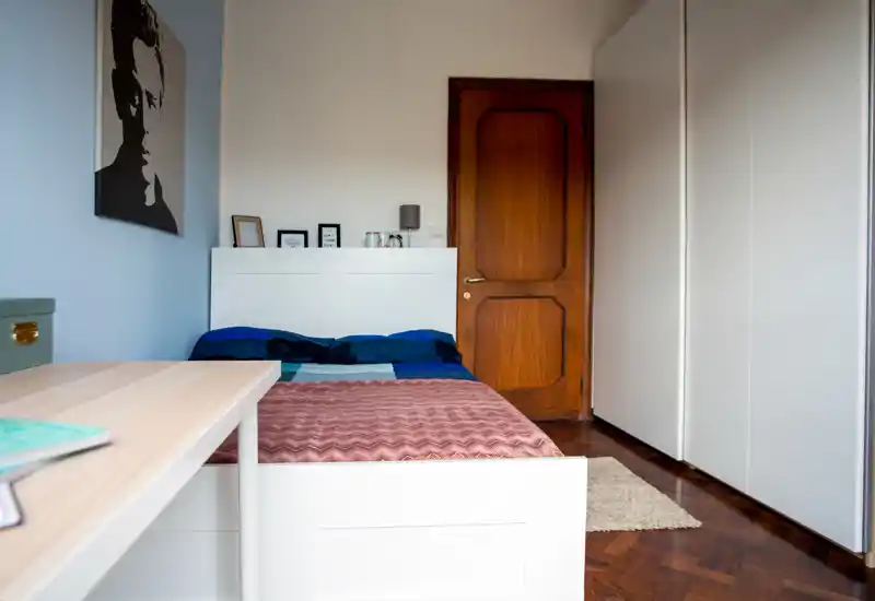 Camera da letto luminosa e ordinata con letto matrimoniale, testiera bianca e ampio armadio; in fondo al letto c’è una scrivania, porta in legno e pavimento in parquet.