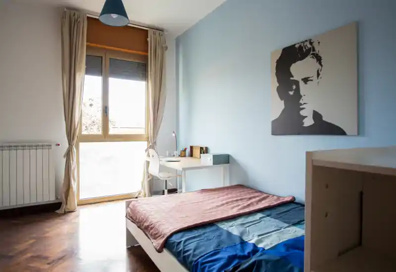 Camera da letto luminosa e minimal con letto singolo, scrivania vicino alla finestra e luce naturale soffusa—ideale per studenti o soggiorni brevi.