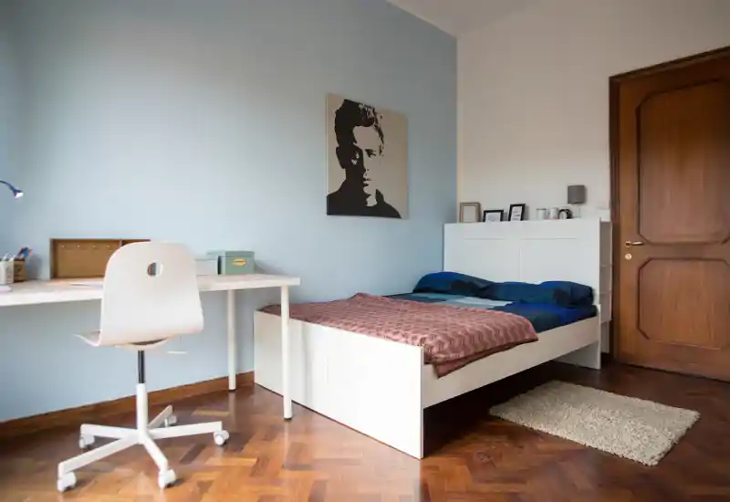 Camera da letto semplice e ordinata con struttura del letto bianca, comodino, scrivania e sedia; parete azzurra e pavimento in parquet creano un'atmosfera luminosa e accogliente.