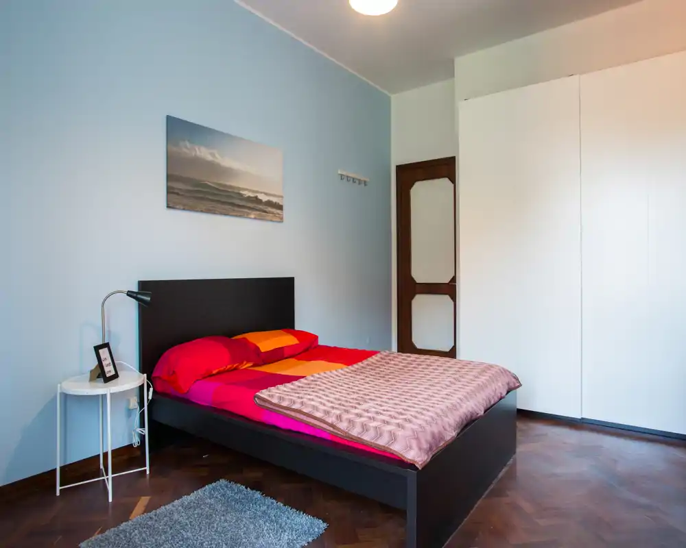 Camera da letto luminosa e ordinata con struttura del letto scura, biancheria colorata e un grande armadio bianco. Il locale presenta una stampa decorativa, comodino con lampada e un tappeto piccolo, che creano un ambiente accogliente.