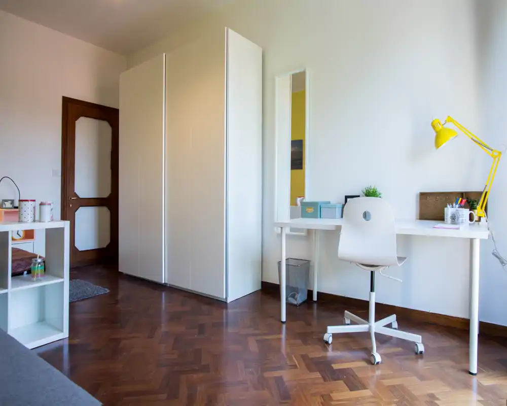 Angolo di camera minimal e ben illuminato con un grande armadio bianco, scrivania e sedia girevole; la zona è ordinata e il pavimento in parquet dona calore.