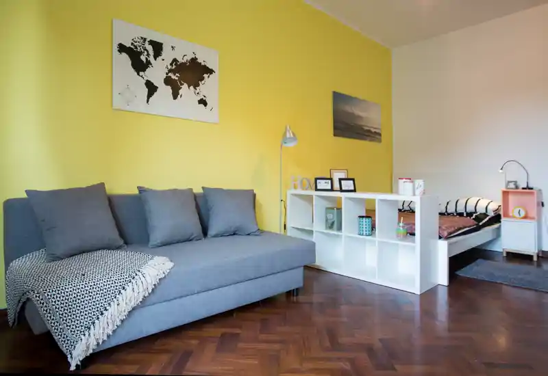 Camera da letto luminosa e moderna con un divano grigio e un letto singolo parzialmente visibile separato da una libreria a cubi bianca; parete gialla e pavimento in parquet creano un ambiente accogliente.