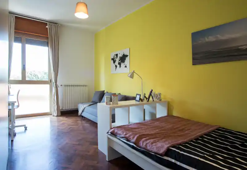 Camera da letto luminosa con letto singolo in primo piano, piccolo divano e scrivania vicino alla finestra; parete gialla e parquet in legno creano un ambiente accogliente e ben curato.