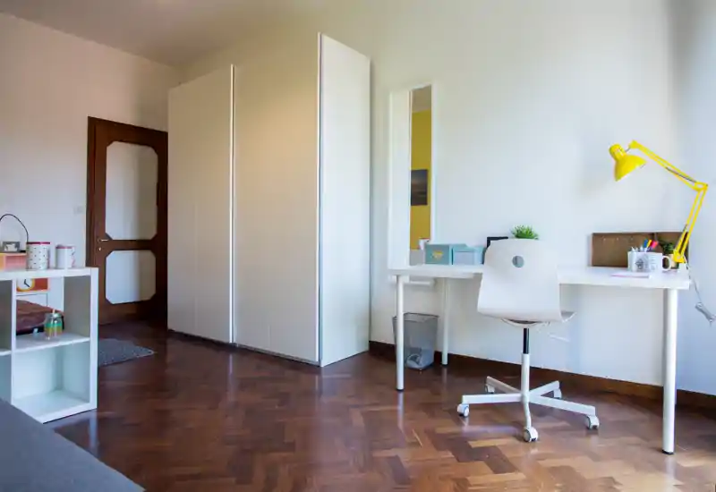 Angolo di camera minimal e ben illuminato con un grande armadio bianco, scrivania e sedia girevole; la zona è ordinata e il pavimento in parquet dona calore.