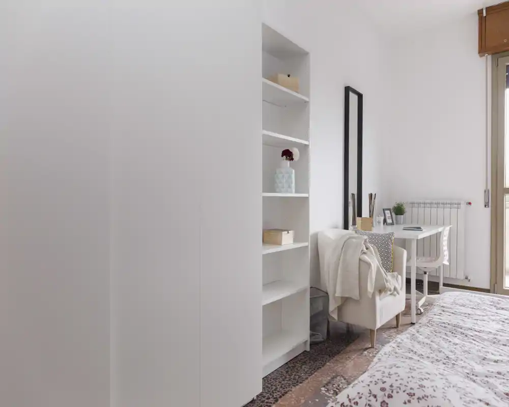 Bright, softly decorated bedroom with a visible double bed, shelving unit, armchair and a small desk by the window — airy, minimal and inviting.