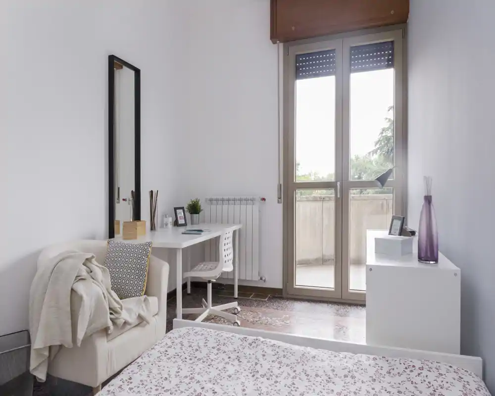 Bright, softly styled bedroom with a double bed in the foreground, a comfortable armchair, a small desk by the window and a balcony door letting in natural light.
