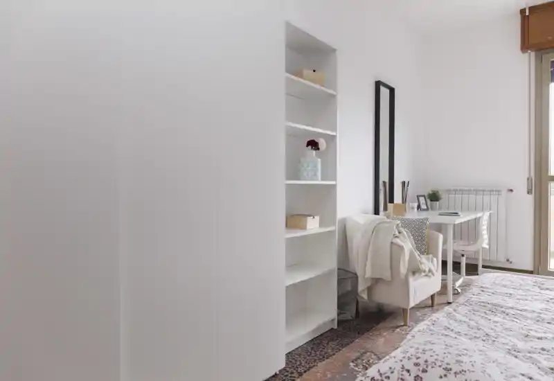 Bright, softly decorated bedroom with a visible double bed, shelving unit, armchair and a small desk by the window — airy, minimal and inviting.