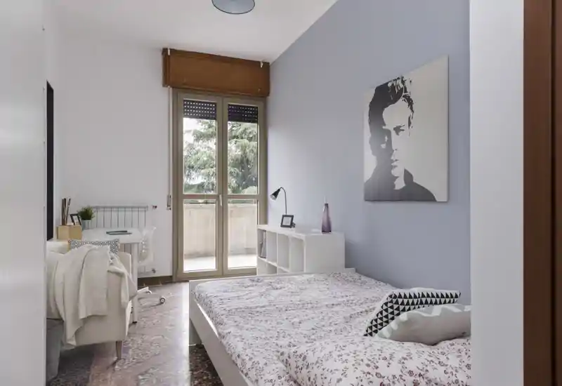 Bright, well-styled bedroom with a double bed facing a balcony door, soft textiles and a minimalist storage shelf beside the bed.