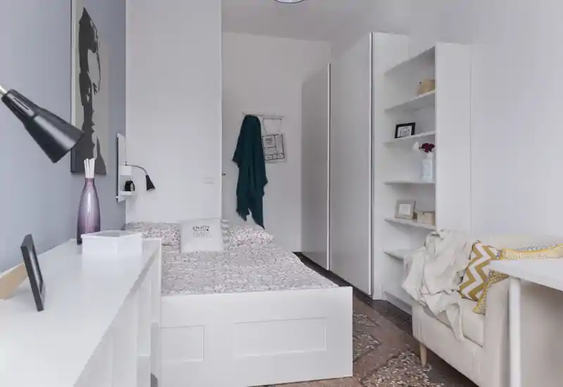 Bright, neatly staged bedroom with a full-size bed, decorative cushions and shelving; clean white furniture and soft textiles create a cozy, modern feel ideal for listings.