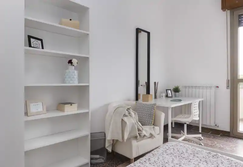 Coin de chambre lumineux et soigné avec un lit confortable, un fauteuil couvert d'une couverture, un bureau près de la fenêtre et des étagères intégrées — parfait pour se reposer et travailler.