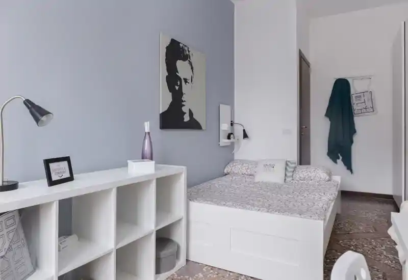 Bright, neatly styled bedroom with a white platform bed, patterned bedding and decorative cushions. The room features minimalist storage shelving, a bedside lamp and calming pastel walls.